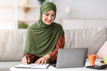 Senior Islamic Businesswoman Taking Notes Sitting At Laptop And Working Online At Home. Muslim Lady Making Financial Report Indoor. Female Business Career And Entrepreneurship, Remote Job Concept