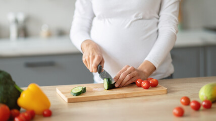 A pregnant woman is chopping cucumber and cherry tomatoes on a cutting board in a contemporary kitchen. Fresh vegetables surround her as she prepares a healthy meal, cropped