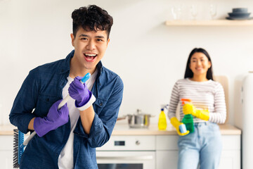 Emotional handsome asian guy imitating rockstar while house-keeping with his pretty girlfriend,...