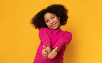 Everything is great. Positive cute little girl gesturing thumbs up and smiling at camera, yellow studio background with empty space