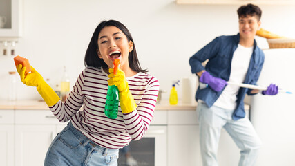 Joyful asian lady with dust cloth using spray as microphone, singing while cleaning kitchen with...