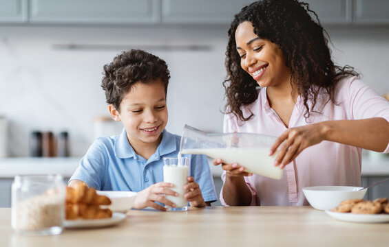 In a cozy kitchen, a mother lovingly pours milk for her son, who smiles with delight. They share a special moment together, surrounded by breakfast items and warmth.