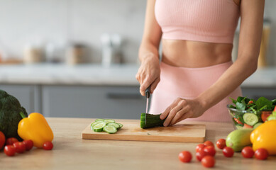 A woman wearing a pink workout outfit skillfully chops cucumbers on a wooden cutting board. Fresh...