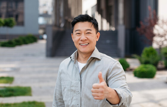Portrait of positive asian man standing outdoors and gesturing thumb up, walking in urban city area, looking and smiling at camera, free copy space
