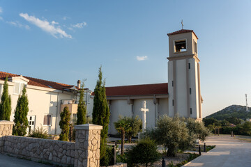 Modern church building with bell tower, white cross monument, and lush garden surrounded by olive trees and tall cypress trees. Concept of peaceful architecture, religious landmarks.