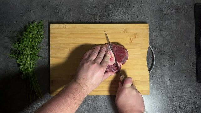 Horizontal 4K video. 60 fps. Top view. Man tries to cut piece of meat on bone with blunt, not sharp knife, beef on wooden cutting board on gray kitchen table. Problem of cooking, lunch, dinner. Dill