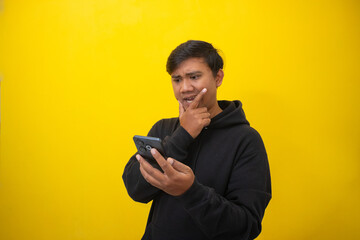 Asian man (Indonesian) positively concentrated at smartphone screen received message dressed in black hoodie isolated yellow wall.