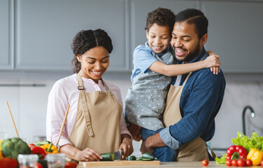 In a cozy kitchen, a father smiles while holding his son, who is playfully seated on his shoulder. The mother, focused on slicing vegetables, shares a moment of affection with her family.