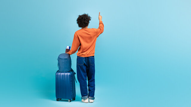A man of African descent is standing against a light blue backdrop, raising his hand as touching invisible screen while waiting near his luggage, copy space - Powered by Adobe