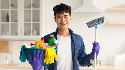 Smiling asian guy ready for cleaning apartment, holding mop and other cleaning tools, white cozy...