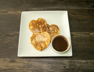 Three heart shaped pancakes with chocolate syrup on the side.