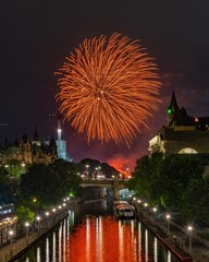 A dramatic display of golden fireworks forms a large, circular burst in the night sky. The glowing reflections on the canal water add warmth and elegance to this striking urban celebration scene.
