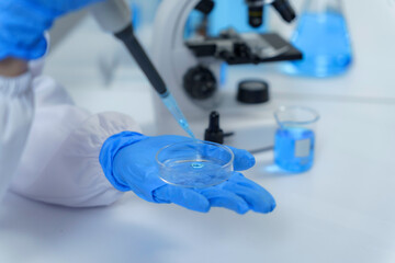 Close up of scientist's hand in blue gloves dropping blue liquid on petri dish using pipette, with microscope and beakers in background