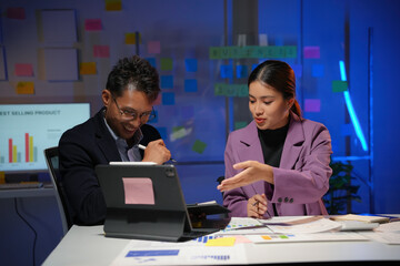 Two businesspeople reviewing financial charts and working together on a digital tablet in a modern office at night