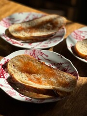 Delicious bread with fruit jam served on delicate red-designed plates, illuminated by warm sunlight. A cozy and inviting breakfast or tea-time setting emphasizing simple and rustic foods.