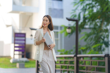 Fototapeta premium Young asian businesswoman holding a laptop while walking through a city park, gazing thoughtfully into the distance