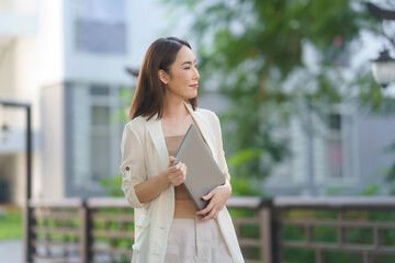 Young asian businesswoman holding laptop walking in urban park looking away, dreaming of future success
