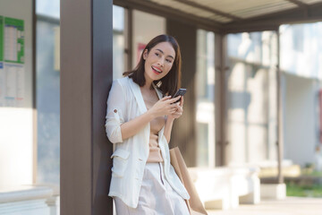 Happy businesswoman waiting at the bus stop, using her smartphone while holding a shopping bag, enjoying urban life and convenience