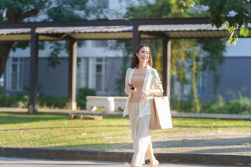 Young business woman is walking in a park, holding a shopping bag and a smartphone, enjoying the sunny day after a successful shopping trip