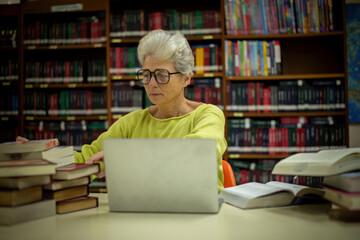 Elderly woman sitting in a library, combining traditional and modern approaches to learning or research by book and laptop computer sense of purpose dedication to personal grown concept.