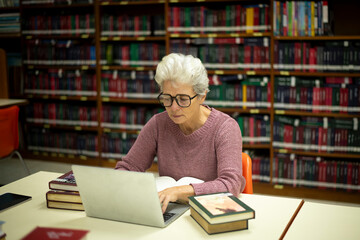 Elderly woman sitting in a library, combining traditional and modern approaches to learning or research by book and laptop computer sense of purpose dedication to personal grown concept.