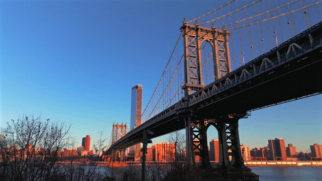A low angle aerial view of the Manhattan Bridge, Brooklyn side with Manhattan in the background. Shot during a golden sunrise. The camera dolly in and boom up, tilted up towards one of the towers.