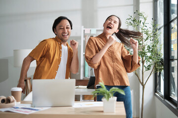 Happy working in casual small business office. Two Asian startup company colleagues enjoy dancing and singing songs together, relaxing from freelance jobs, humor, and funny coworker team in workplace.