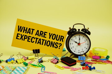What Are Your Expectations? text on paper card with alarm clock and stationery on yellow background