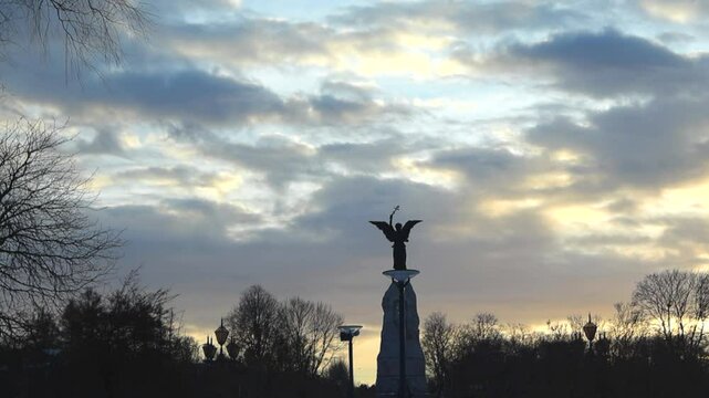 Moving footage of Russalka monument during sunset with gray and orange clouds in the sky. The statue is holding up an Orthodox cross to the sky with trees and plants in the background as silhouettes.
