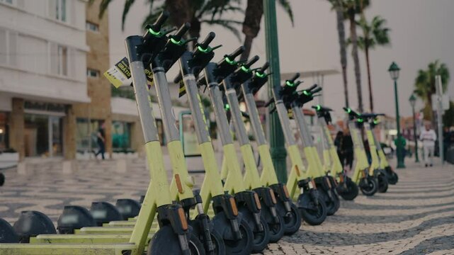 A row of electric scooters parked on a patterned city street, with palm trees and modern buildings in the background. Urban, eco-friendly transportation concept. Electric Scooters Lined Up on City