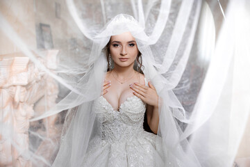 A woman in a white wedding dress is standing in front of a white curtain. She is wearing a veil and...