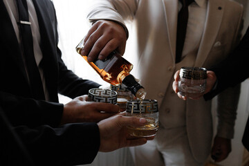 A man is pouring a drink into a glass while another man watches. The scene is set in a bar or a social gathering where people are enjoying drinks and each other's company. The atmosphere is relaxed