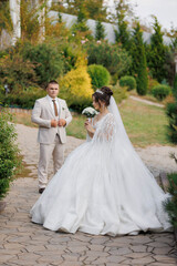 A bride and groom are standing in front of a green hedge. The bride is wearing a white dress and holding a bouquet. The groom is wearing a suit and tie. The scene is set in a garden
