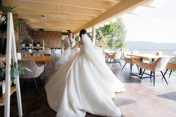 A bride and groom are standing in front of a restaurant with a view of the mountains. The bride is wearing a long white dress and the groom is wearing a suit. The scene is romantic and picturesque