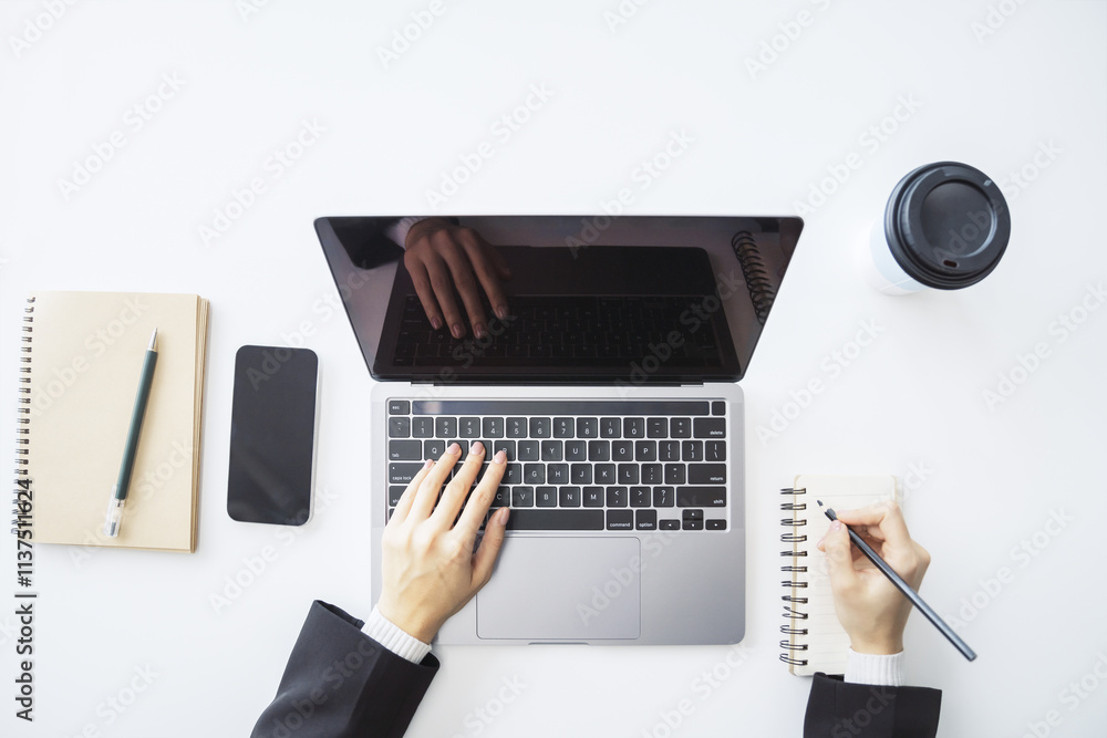 Poster detailed view of a woman's hand jotting down notes in a notepad on a modern laptop, with a blurred b
