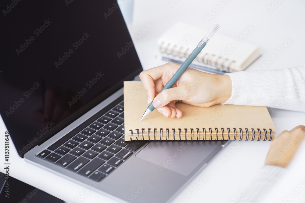 Poster close-up capture of a woman's hand as she writes in a notepad on a sleek laptop, with an out-of-focu