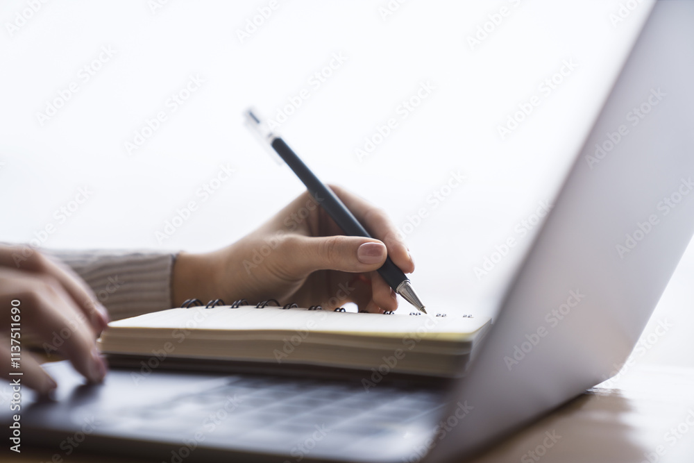 Poster close perspective of a woman's hand making entries in a notepad on a cutting-edge laptop, with a sof