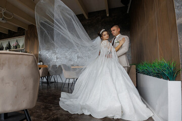 A bride and groom are standing in front of a table with chairs. The bride is wearing a white dress...