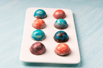 Assorted multi-colored homemade chocolates. Modern hand-painted chocolates on a white plate on a blue background. Product concept for chocolatier. Selected focus