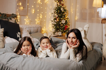 mother and two children lying on bed on christmas happy evening in cozy room decorated with garlands