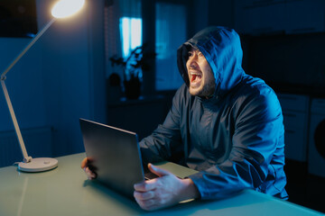 Cyber failure depicted by angry hacker at table in dimly lit home.