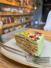 A slice of layered pistachio cake with green frosting and edible rose petals, served on a rustic plate in a cozy, warmly lit caf during daytime