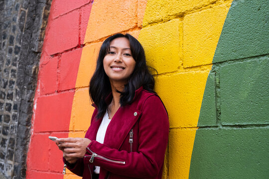 Smiling woman leaning with mobile phone on rainbow wall