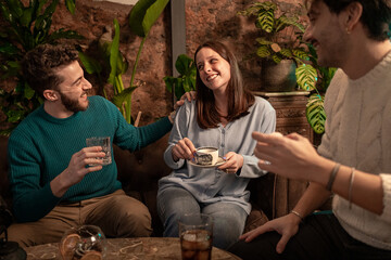 Young friends sharing drinks and laughter at cozy cafe with tropical decor - casual evening...