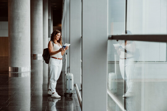 Mature woman examining passport and standing with suitcase near window