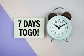 7 Days To Go! text on paper card with alarm clock on pink and purple geometric background