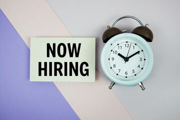 Now Hiring text on paper card with alarm clock on purple and pink geometric background