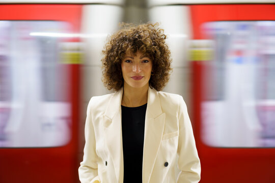 Smiling curly haired businesswoman standing in front of moving train at station