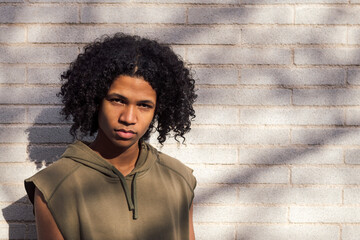 african american male teen with afro hairstyle wearing casual clothes standing looking at camera in sunlight against white brick wall, concept of youth and urban lifestyle, copy space for text