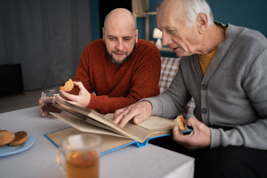 An elderly father and his adult son look at a photo album, remembering happy moments from the life of a young man while having fun together at home